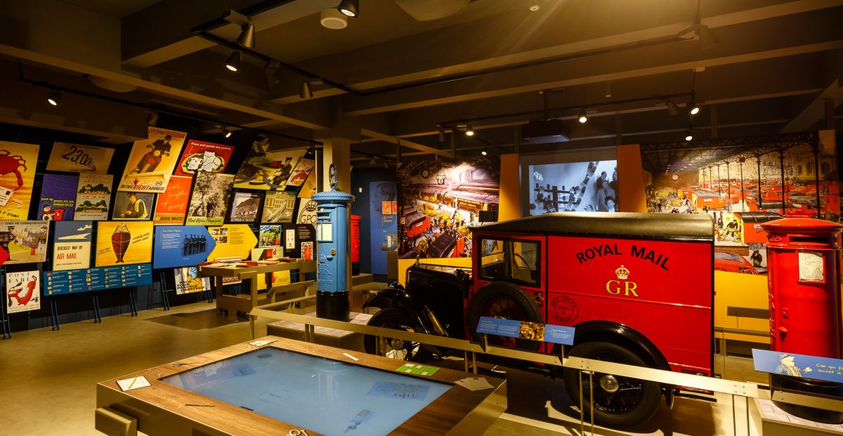 The exhibition space inside the postal museum can be seen. In the shot you can see a blue postbox, lots of old posters and a bright red postal van that was used many years ago.