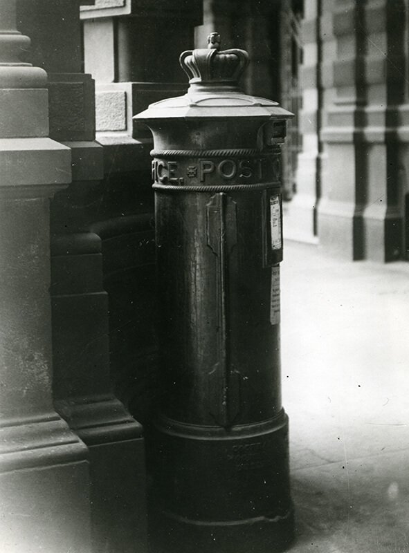 A pillar box for letters standing on the corner of a street, with a metal crown attached to the top of the box. The words 'Post Office' are on the pillar of the box.