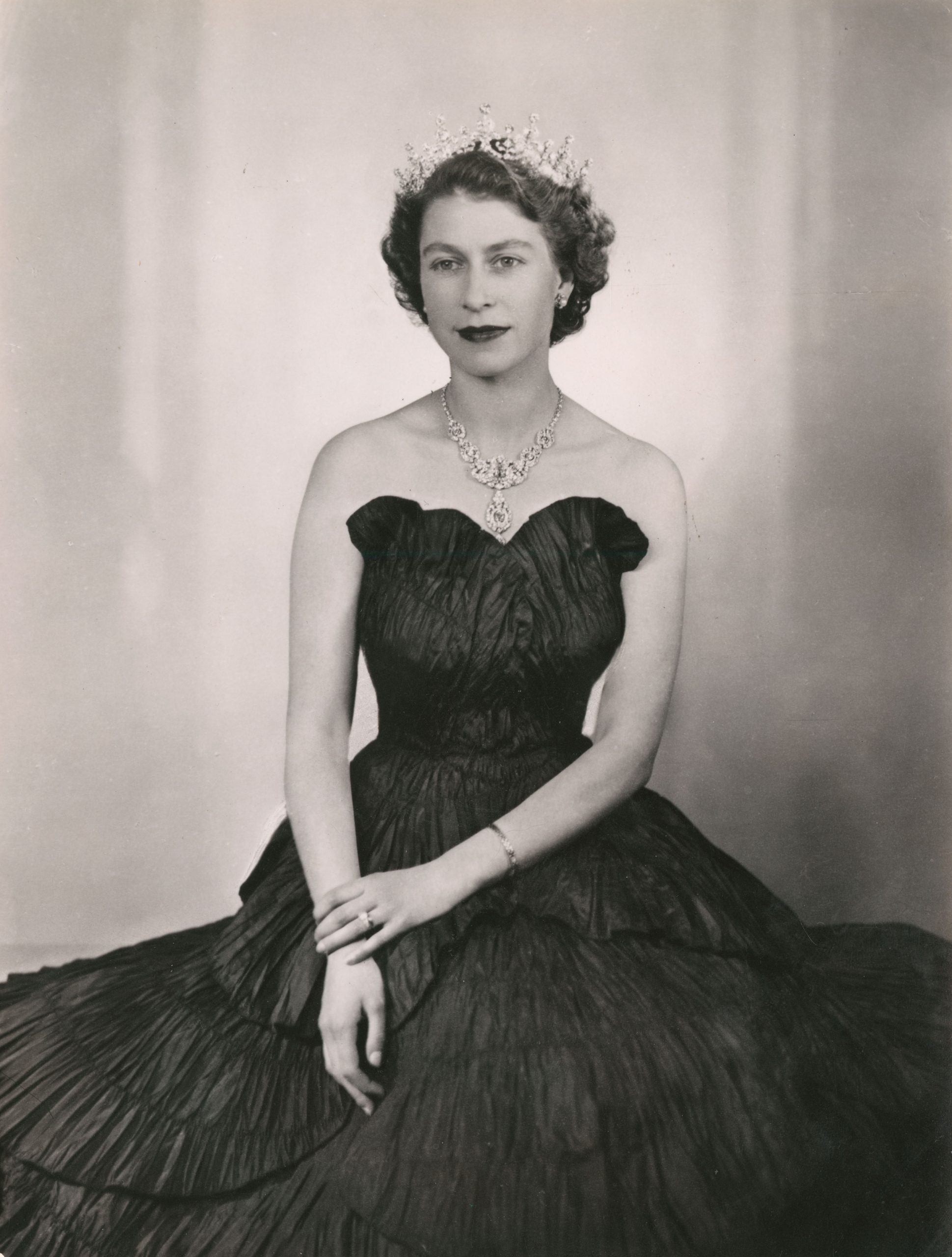 Black and white image of the Queen in a black ruffled dress wearing a tiara. 