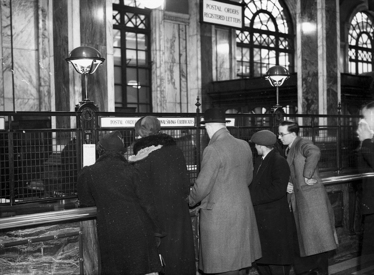 Black and white photograph of five people standing at a post office counter. The photograph is taken from behind the customers. The counter is wood with a metal divider. There is a sign with a white background and black text for postal orders and national savings certificates.