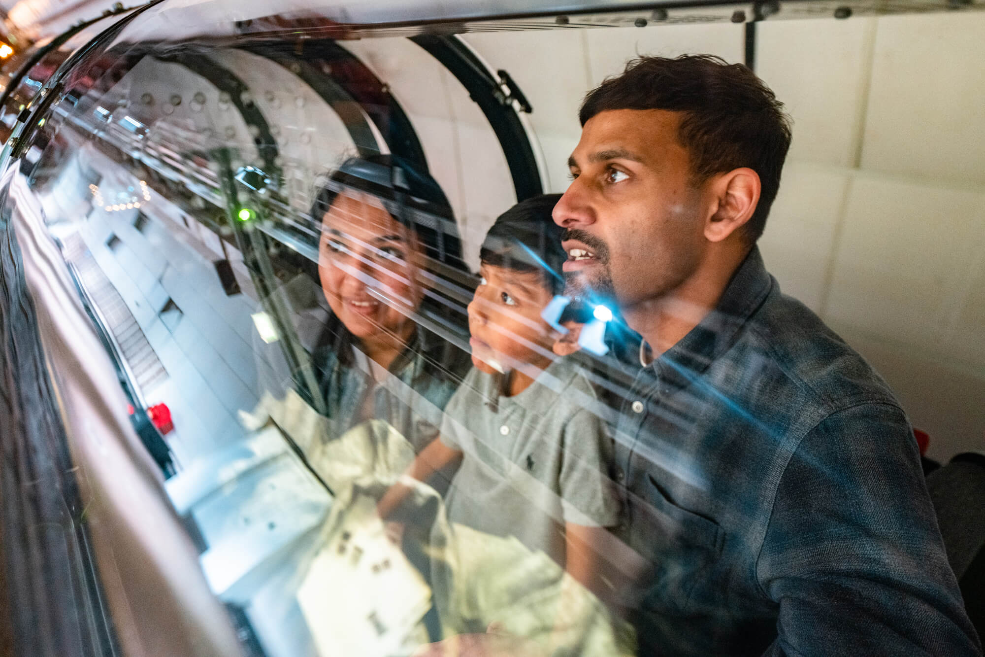A Mum and Dad sit on Mail Rail with their son. All three people have brown skin and dark hair. They sit watching the audio-visual part of the ride.