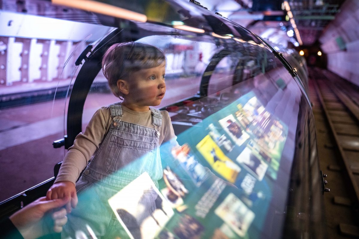 A young child stands up inside a Mail Rail carriage. He looks in awe at the visuals that are playing on the platform screen. 